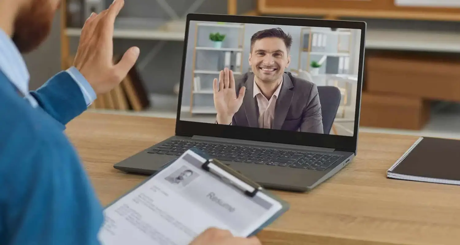 HR professional holding a candidate resume during a video call, with a smiling male job applicant waving from the laptop screen on a wooden desk.
