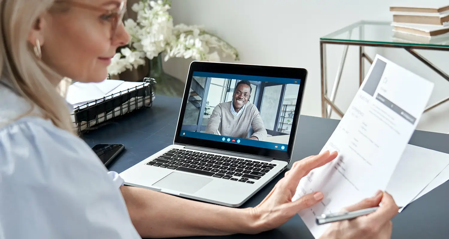 Hiring manager reviewing a printed resume document while conducting a remote video interview with a smiling male candidate on a laptop screen.