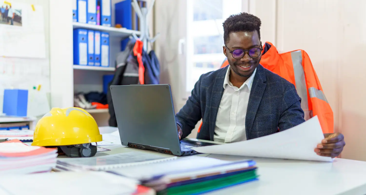 Construction admin in glasses reviewing project documents at a desk with a yellow hard hat and laptop nearby.
