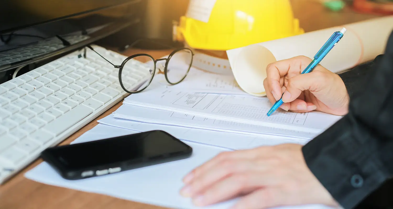 Hands writing on construction blueprints at a desk with a hard hat, glasses, keyboard, and phone.