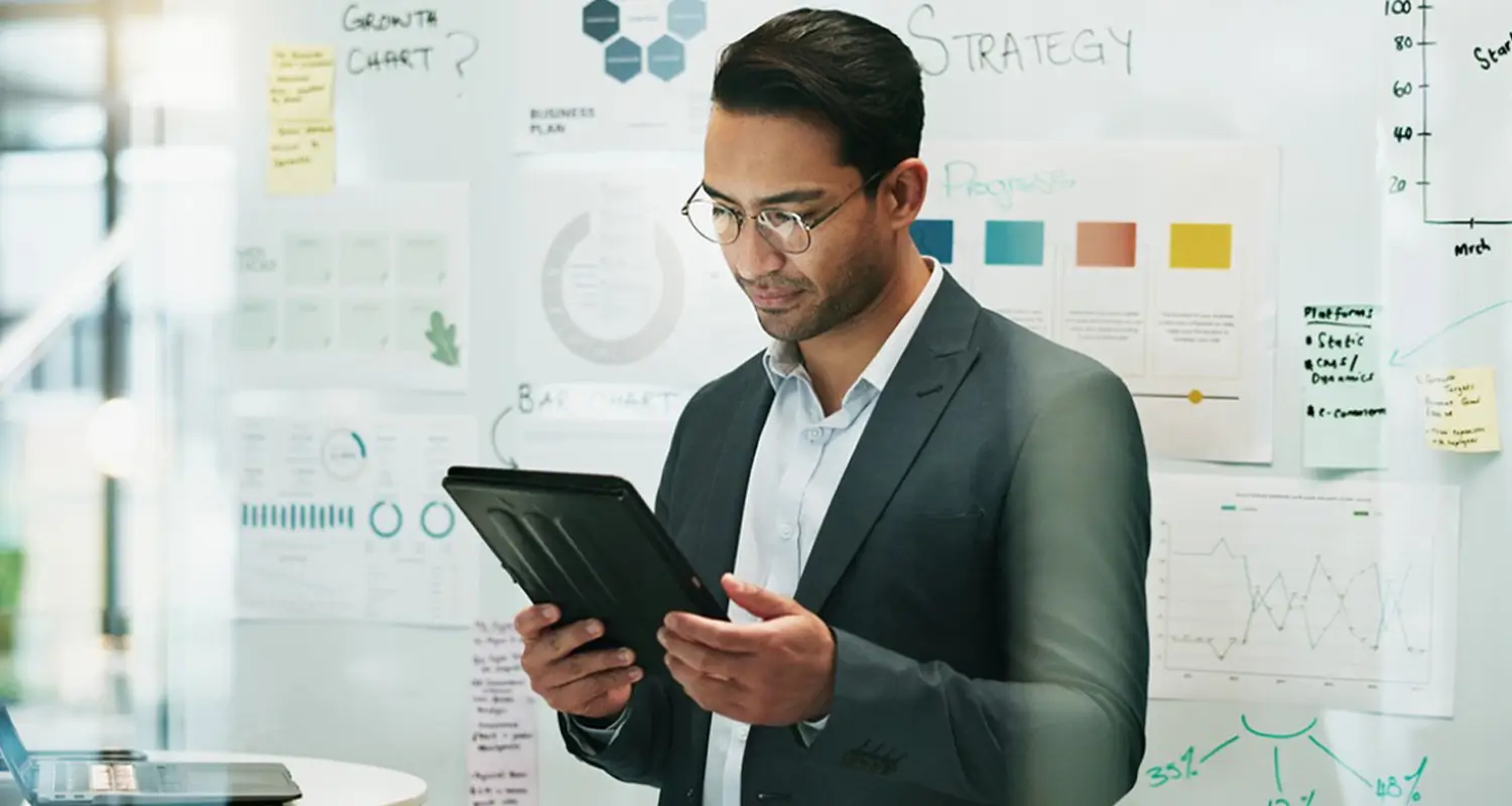 Business analyst in a suit reviewing a tablet with strategy plans and financial charts on a whiteboard behind him