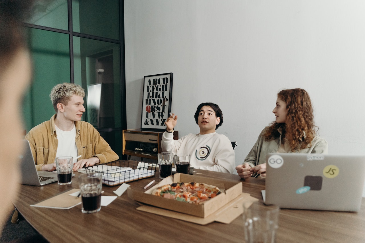 Three colleagues sitting around an office table with laptops, drinks, and an open pizza box, engaged in casual conversation. The setting suggests a relaxed team lunch or informal meeting, illustrating employee perks and team culture benefits.