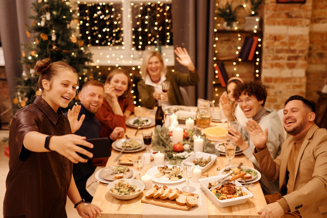 A group of seven people enjoying a festive holiday dinner around a decorated table with candles, food, and drinks. One person takes a selfie as the group waves and laughs, representing a team celebration or Christmas bonus occasion.