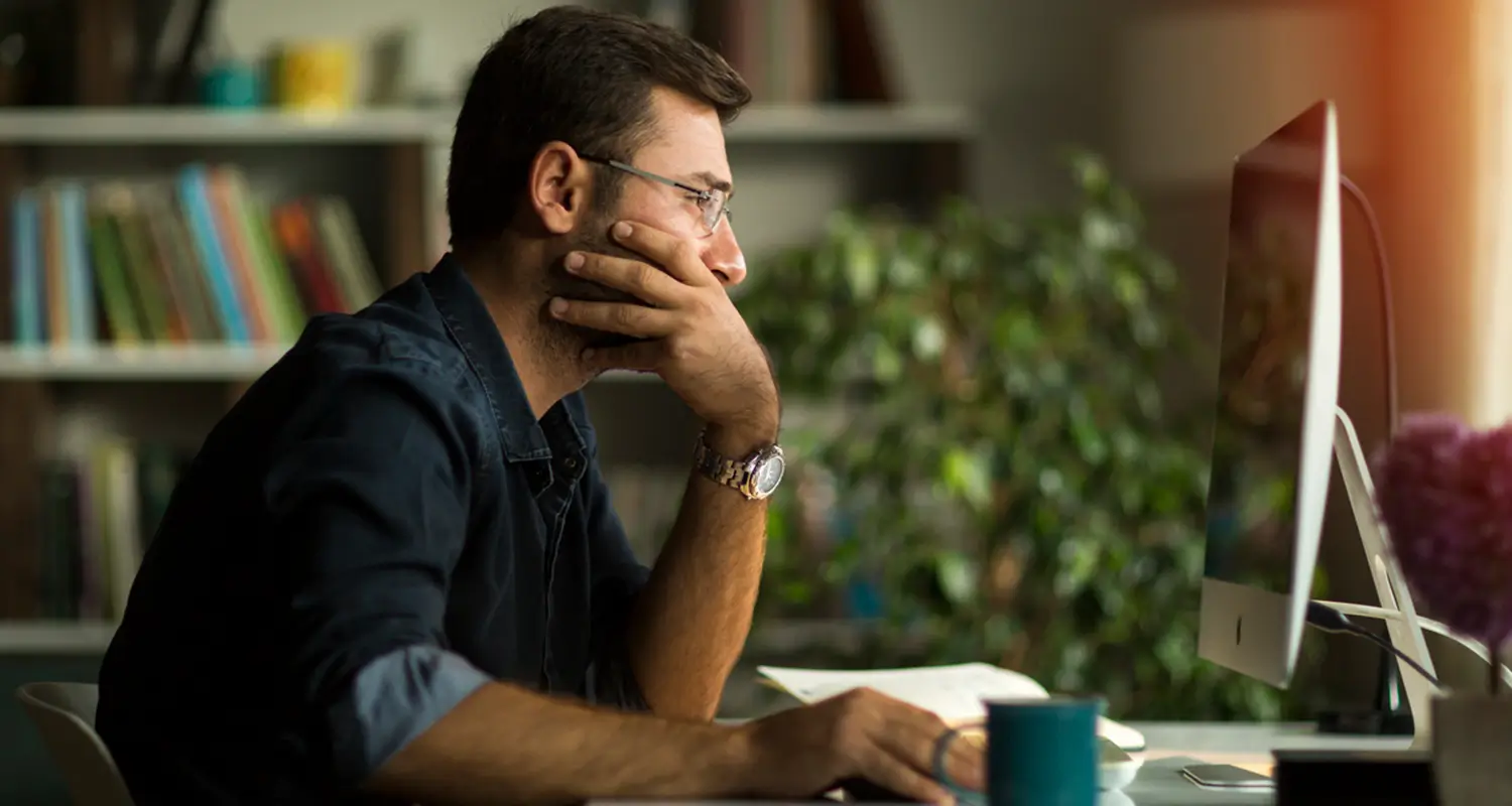 A man with glasses sits at a desk in a home office, resting his chin on his hand while staring intently at an iMac monitor. A blue coffee mug, an open notebook, and a bookshelf with plants in the background suggest a focused, contemplative work session.