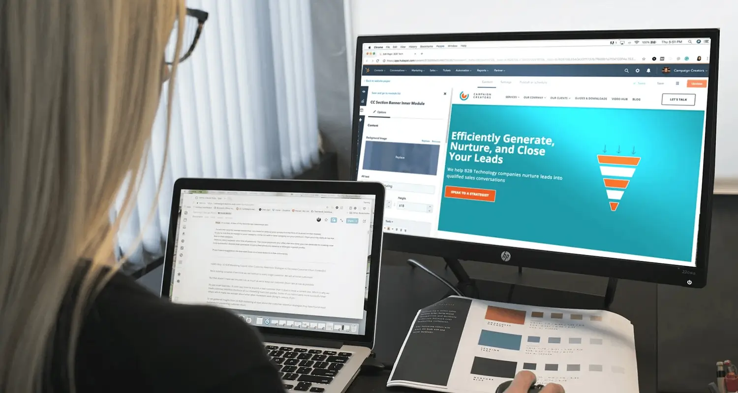 A woman with blonde hair works at a dual-screen setup, with a laptop on the left and an HP monitor on the right displaying a HubSpot CMS page editor alongside a Campaign Creators website with the headline "Efficiently Generate, Nurture, and Close Your Leads." A printed color-swatch brochure sits on the desk beside her.