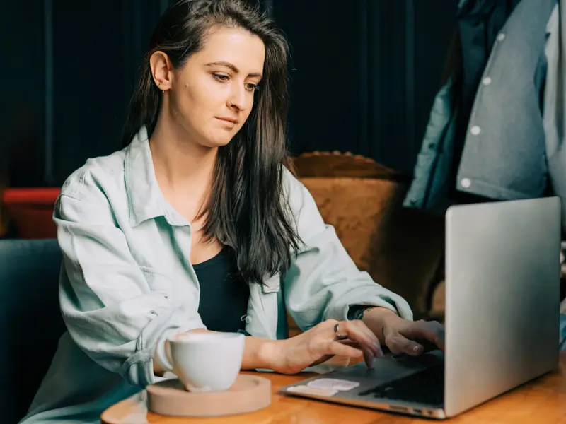 A young woman with long dark hair wearing a light teal button-up shirt sits at a wooden table in a cozy, dimly lit setting, focused on typing on a silver laptop. A white coffee mug on a wooden coaster sits beside her, suggesting a remote worker or talent acquisition professional working from a café or home office.