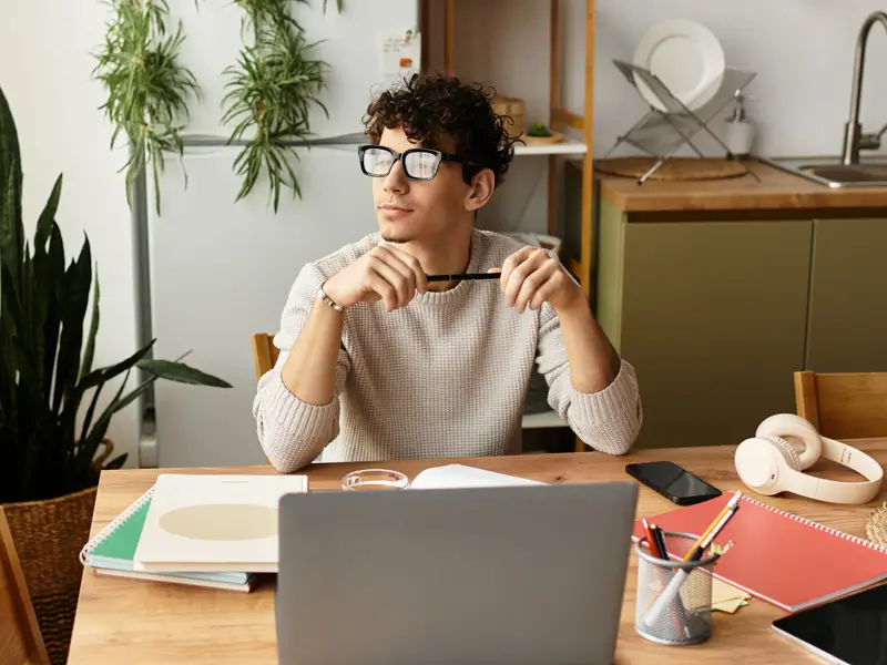 A young man with curly dark hair and black-framed glasses sits at a wooden home-office desk, holding his glasses thoughtfully while gazing away from his laptop. Open notebooks, a smartphone, headphones, and pens are spread across the desk, and lush houseplants fill the background, evoking a reflective moment during a remote job search or hiring process.