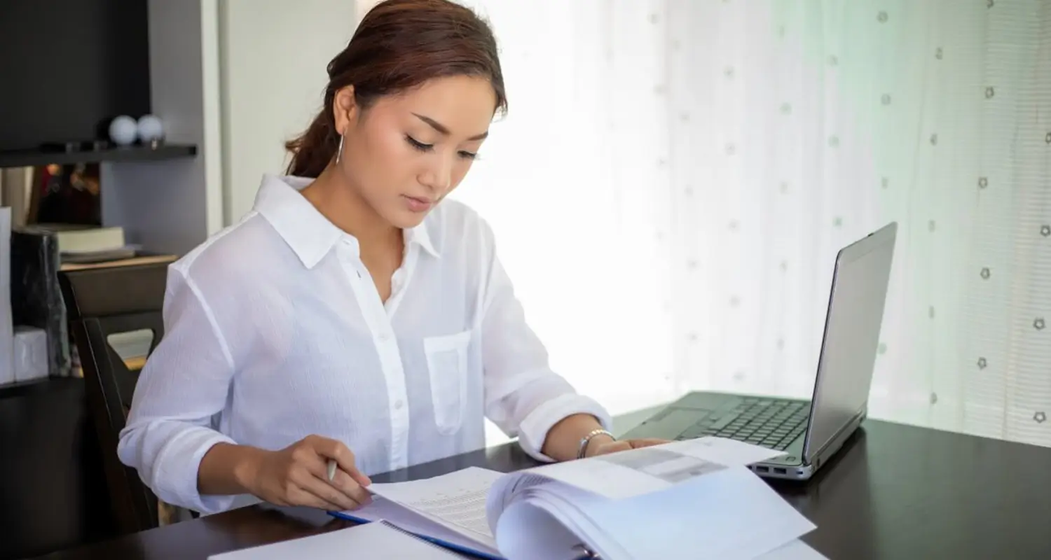 A woman in a white button-up shirt sits at a dark desk reviewing printed documents beside an open laptop, with natural light coming through sheer curtains in the background.