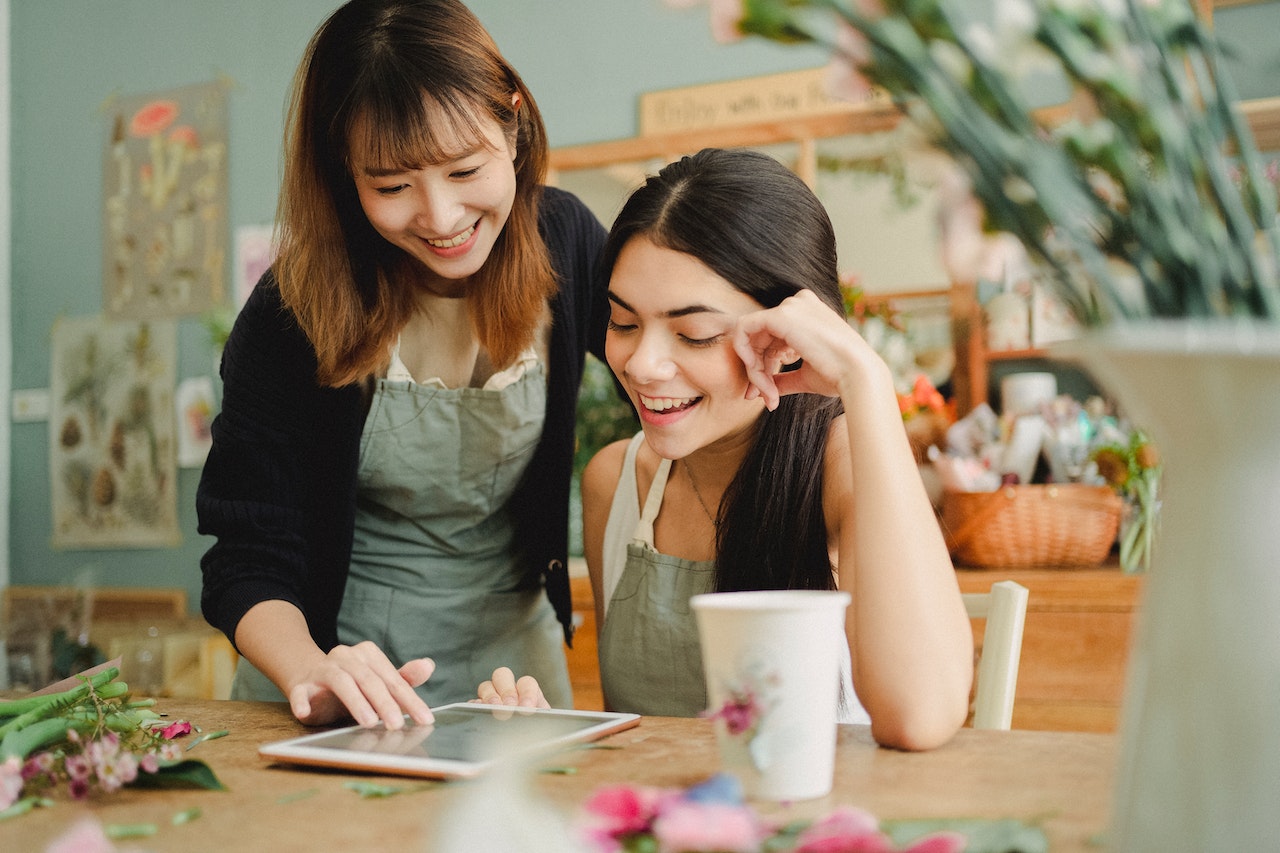 Two women smiling and looking at a tablet together in a flower shop, one pointing at the screen while wearing aprons