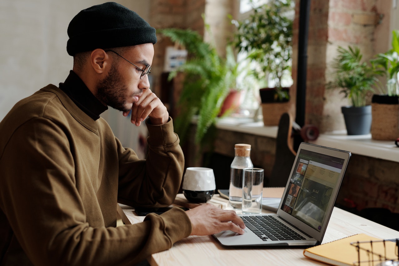 A focused Black man wearing a beanie and glasses sits at a laptop in a plant-filled cafe or creative studio with exposed brick walls and large windows. He is working on design content visible on the screen, with a coffee cup and water bottle nearby, depicting a remote multimedia designer at work.