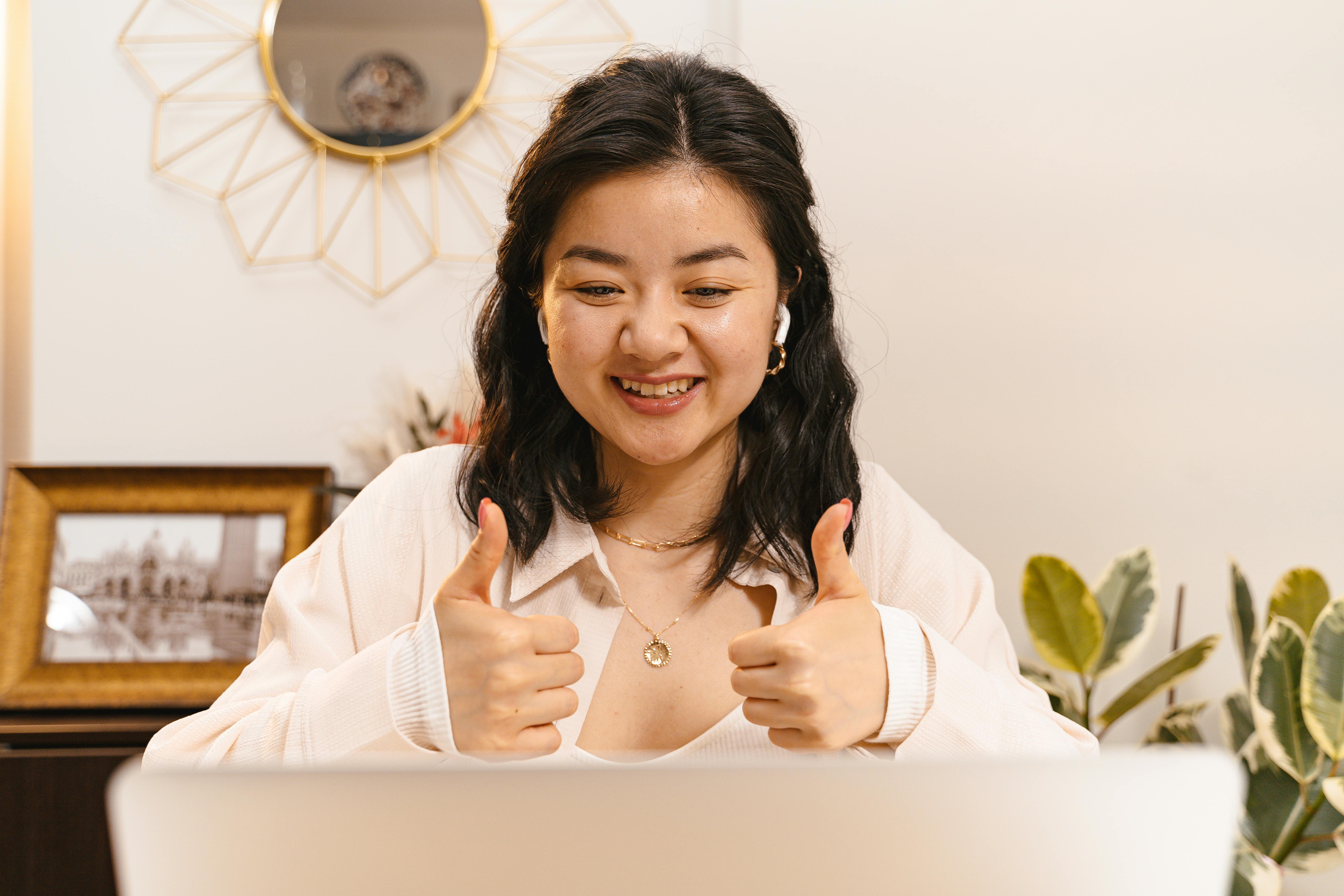 A smiling young woman with dark hair gives two thumbs up while sitting at a laptop, wearing AirPods and a light blouse. A decorative gold sunburst mirror and houseplants are visible in the background, conveying enthusiasm and positive feedback in a remote work setting.