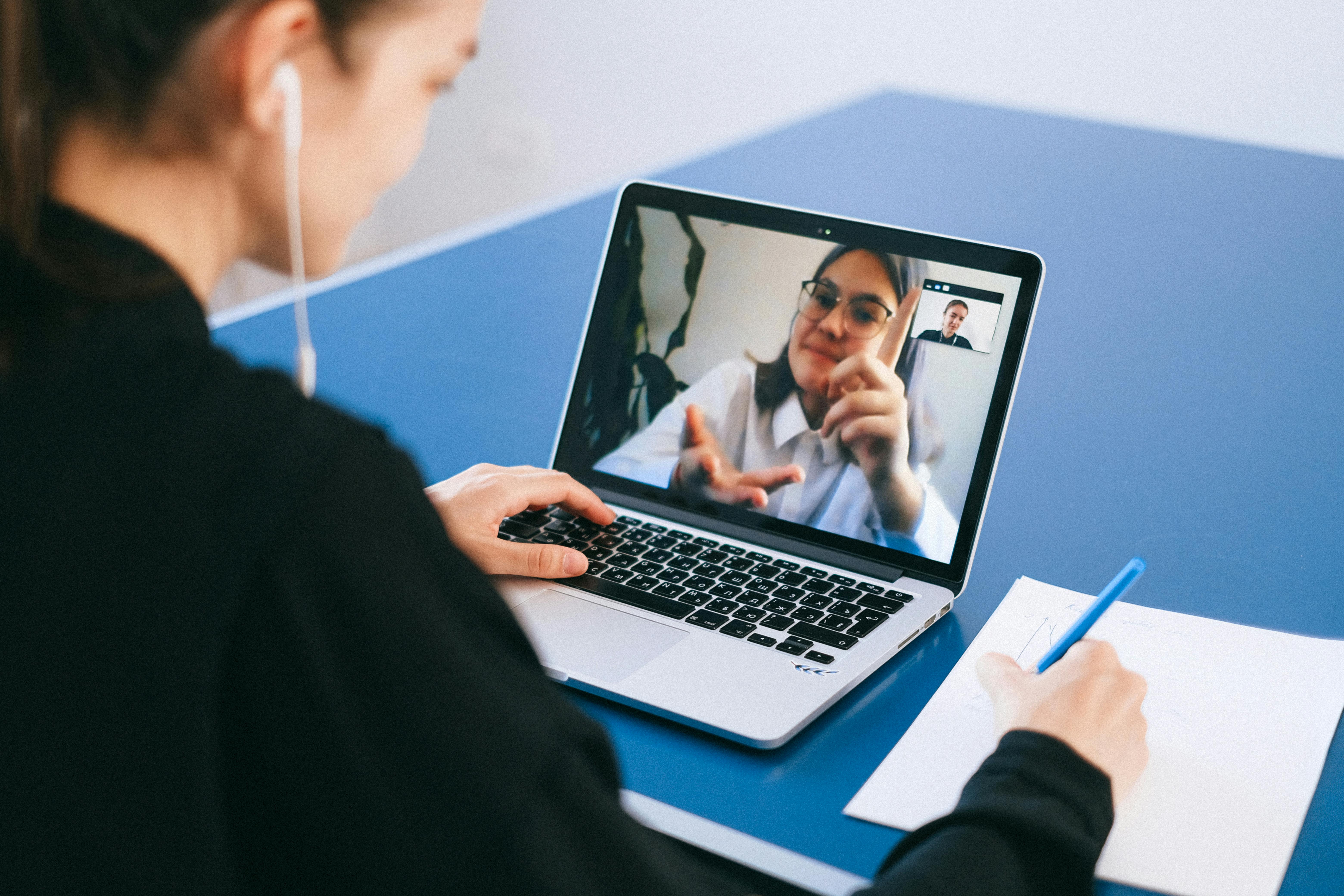 A woman wearing earbuds takes notes on paper while participating in a video call on her laptop. The laptop screen shows a woman in glasses gesturing expressively during the call, with a small self-view thumbnail visible, representing a remote sales meeting or coaching session.