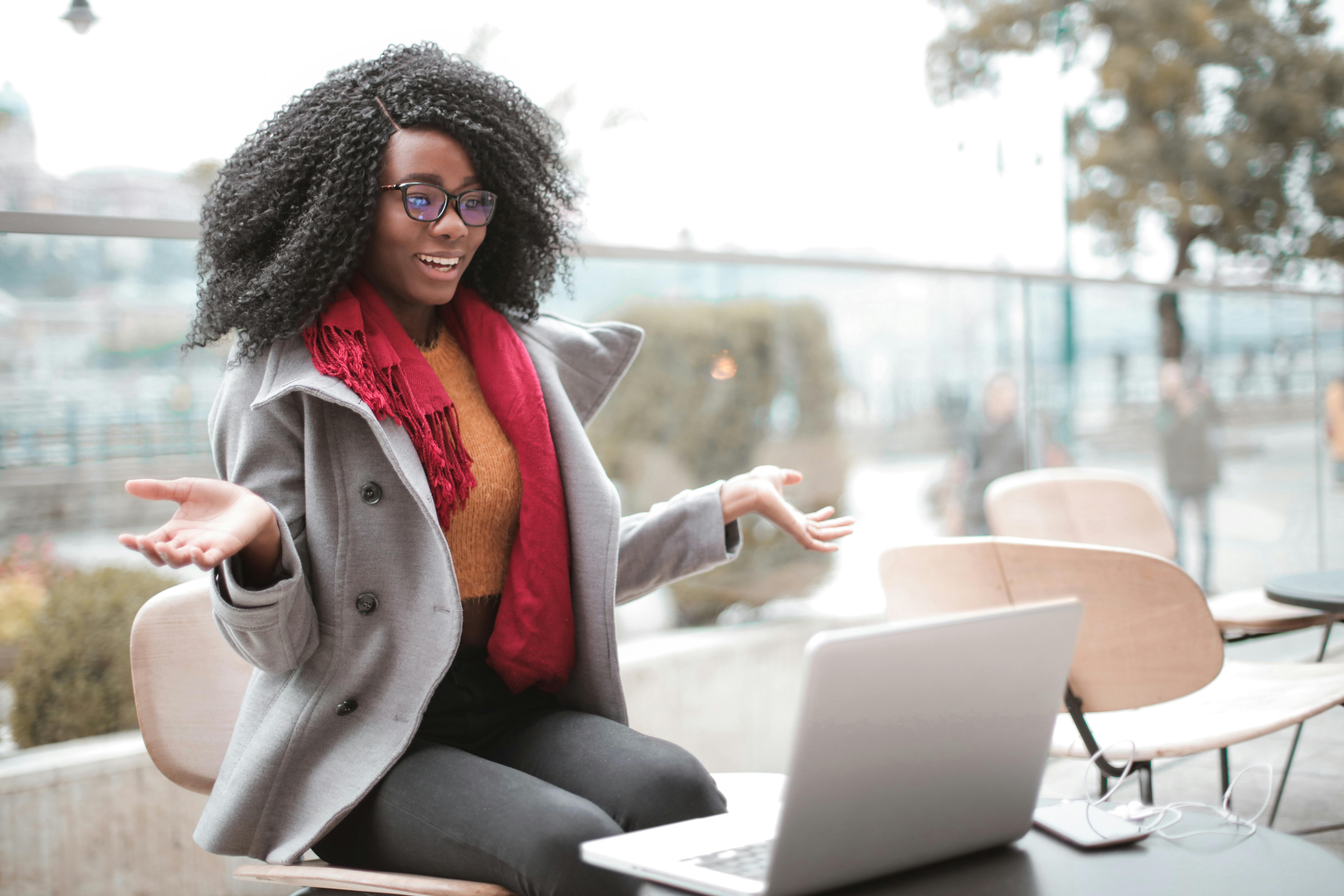 A Black woman with curly hair, wearing glasses, a gray coat, and a red scarf, sits at an outdoor café table with a laptop open in front of her. She gestures with both hands open wide, appearing engaged and expressive, conveying enthusiasm during a remote work session.