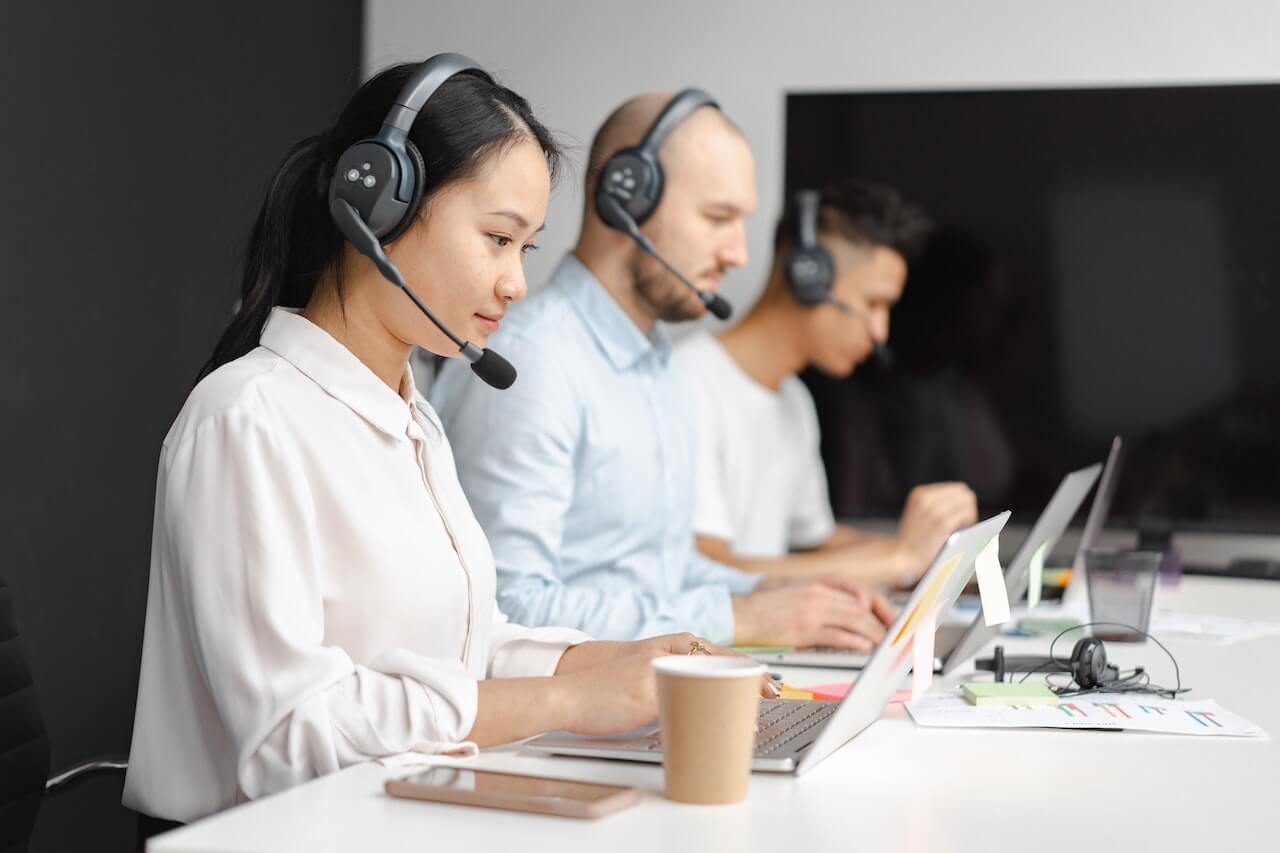 Three customer support agents wearing headsets working on laptops at a shared desk in an office setting