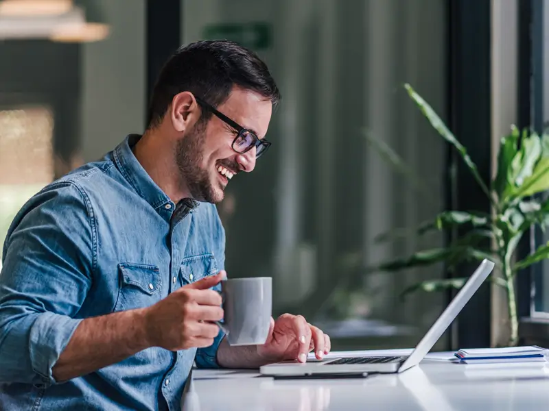 Smiling man with glasses holding a coffee mug while working on a laptop at a modern office desk