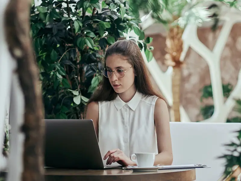 Young woman with glasses working on a laptop at a cafe table surrounded by lush indoor plants