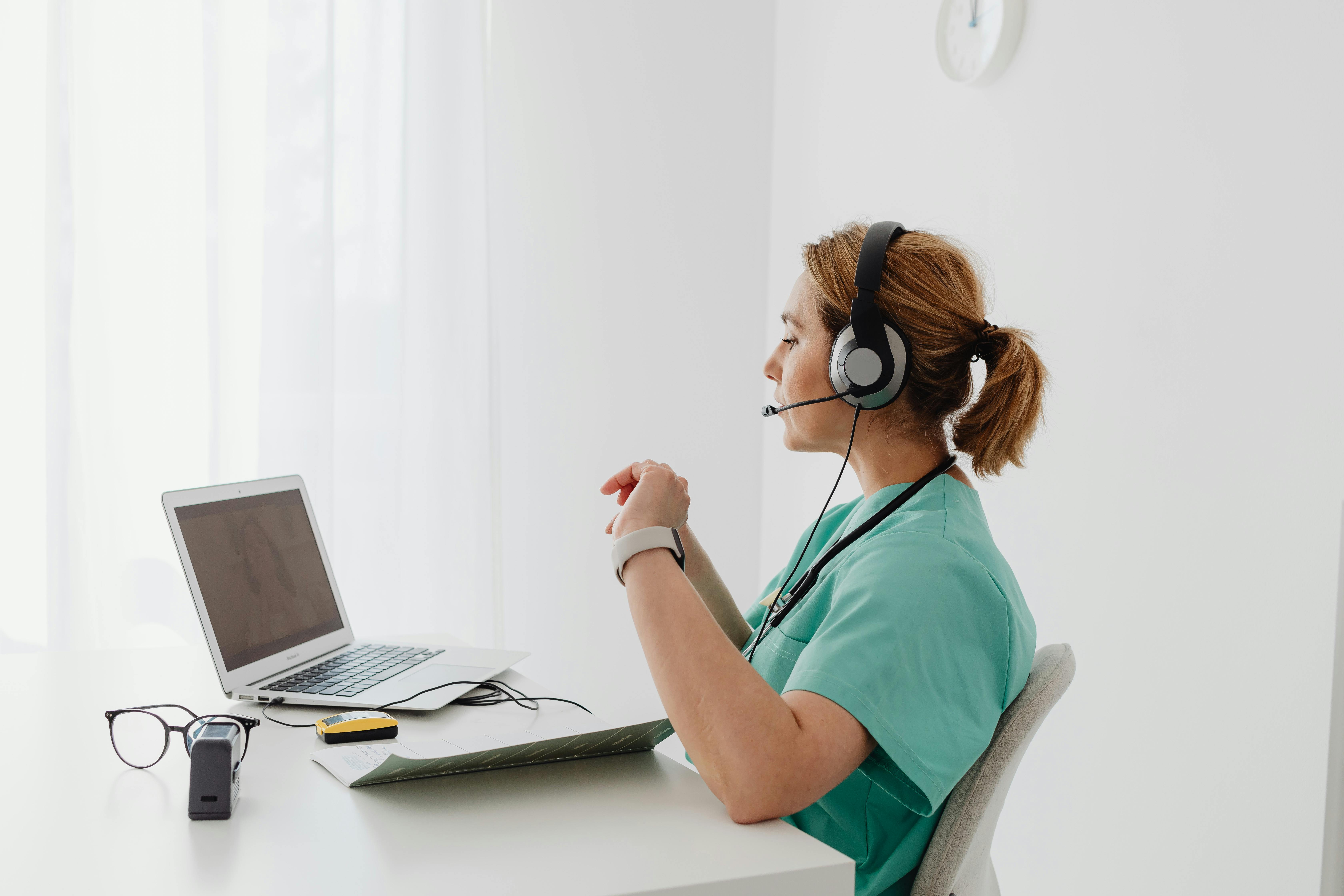 A female healthcare professional in teal scrubs wearing a headset microphone sits at a white desk, looking at a laptop screen with her hands clasped. Eyeglasses, a folder, and other desk items are nearby, depicting a virtual medical assistant handling remote patient communication.