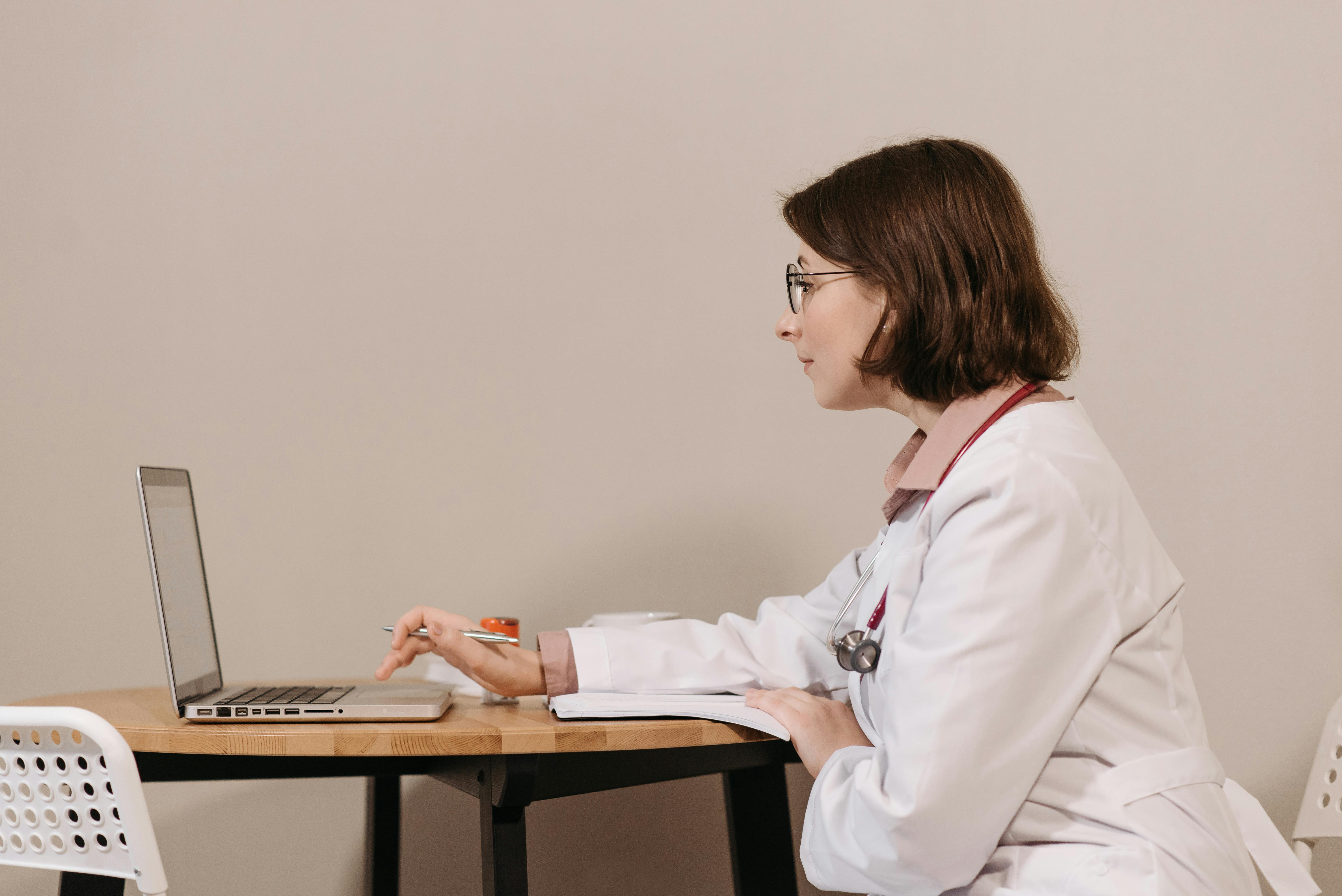 A female doctor wearing glasses and a white lab coat with a red stethoscope sits at a wooden desk, focused on her laptop screen and holding a pen. She appears to be reviewing patient records or administrative tasks in a clean, minimal office setting.