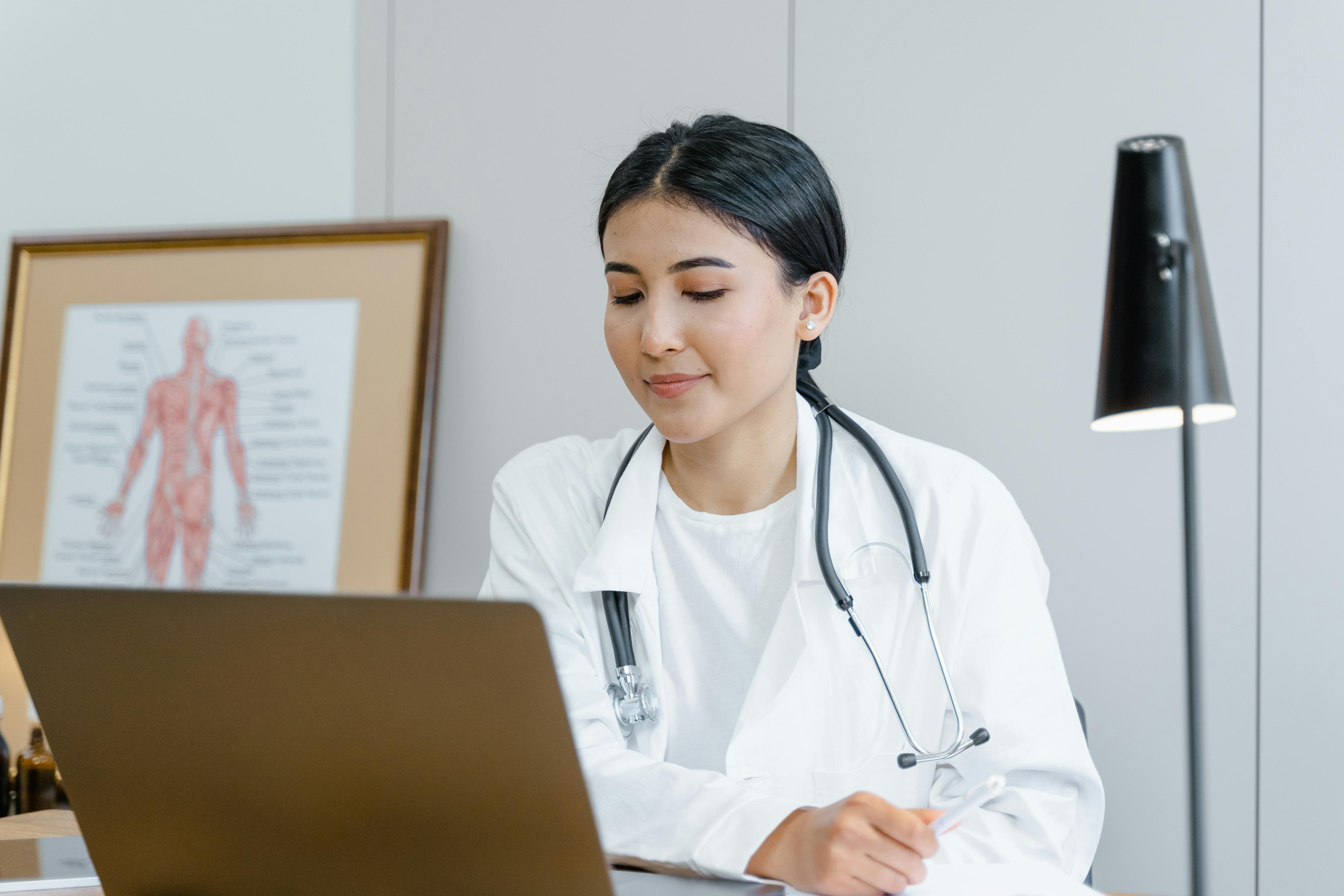 A female doctor in a white coat with a stethoscope smiles gently while working on a laptop at her desk. An anatomical body chart is visible on the wall behind her, and a desk lamp is lit, suggesting she is working in a medical office conducting virtual patient or administrative work.