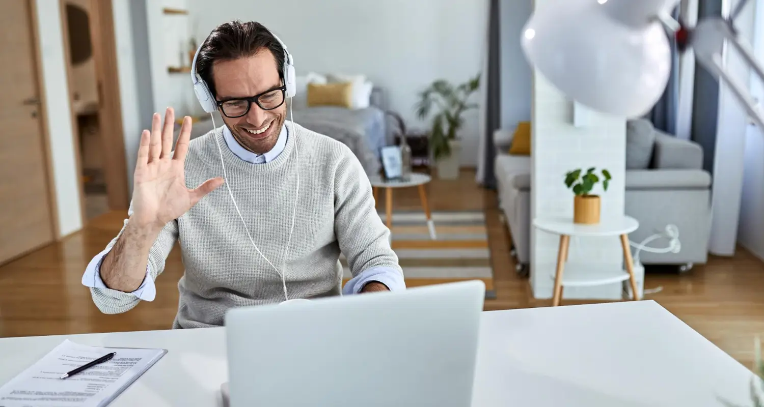 A smiling man wearing glasses and over-ear headphones waves at his laptop during a video call while working from a bright, modern apartment living room. A notepad and pen sit on the desk beside the laptop.
