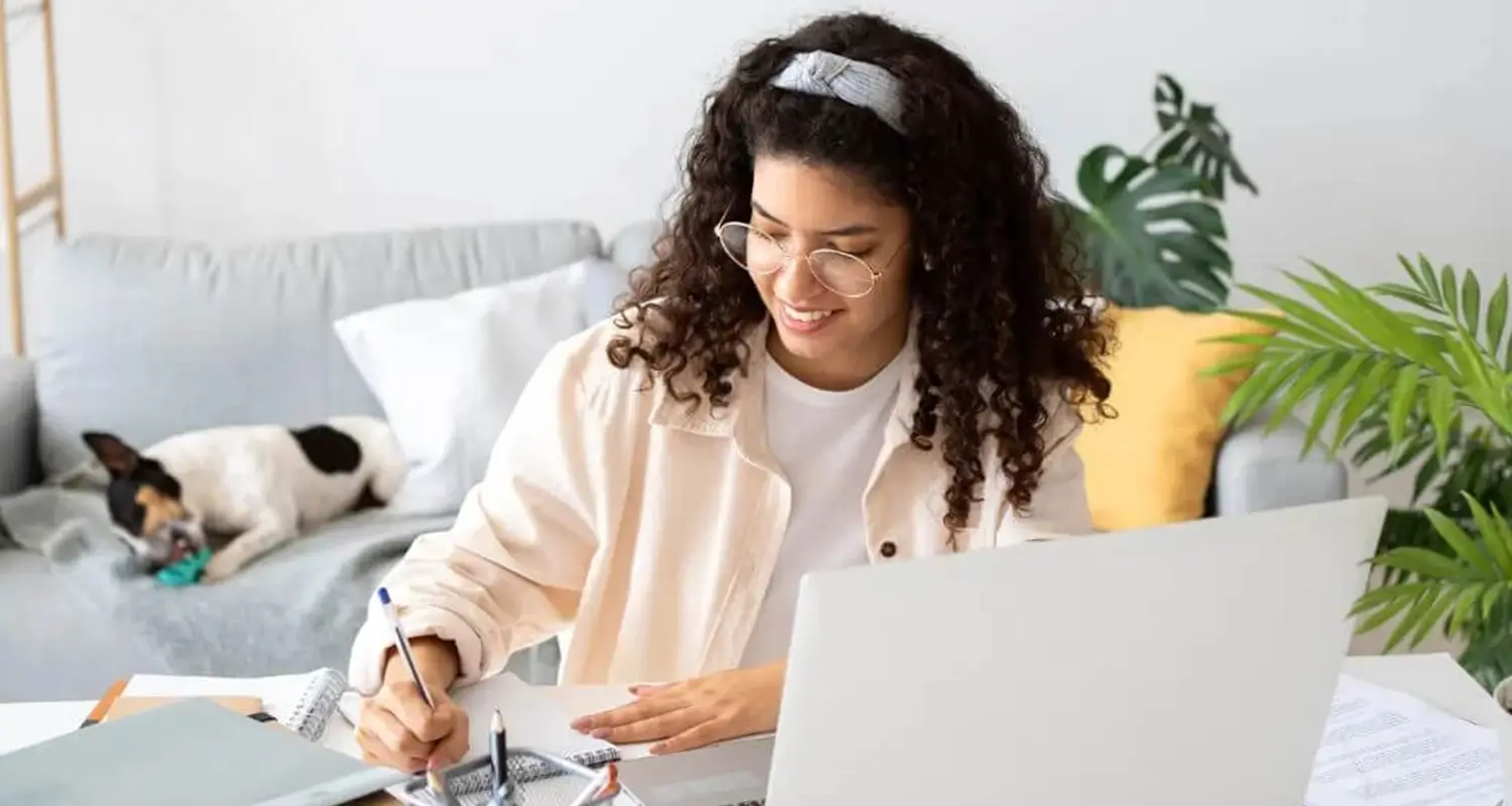 A smiling young woman with curly hair and glasses writes notes in a notebook while working at a laptop from a cozy home setup. A small dog rests on a sofa in the background, surrounded by houseplants and a yellow cushion.