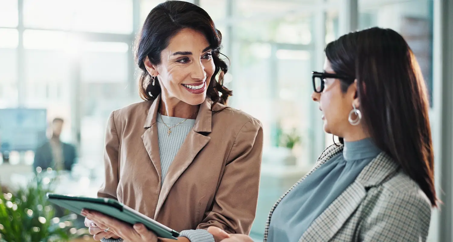 Two professional women stand together in a bright modern office, one in a tan blazer holding a tablet and smiling at the other in a gray blazer and glasses. Their engaged conversation represents the collaborative dynamic between CFOs and their offshore finance team members who drive accounting efficiency and business growth.