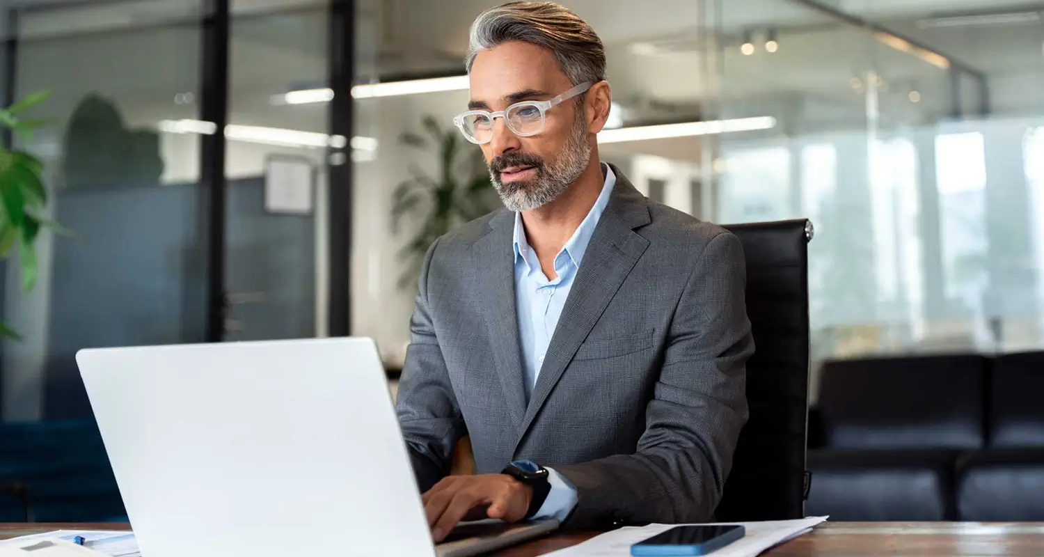 A senior male executive with silver hair and a gray suit sits in a boardroom, focused on his laptop. A smartphone rests on the desk beside him and glass office partitions are visible in the background, representing a CFO or finance leader evaluating the benefits of building an offshore accounting team.