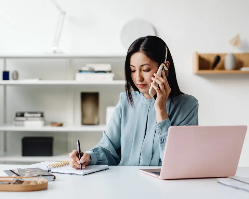 A woman with dark straight hair in a light teal blouse sits at a clean, minimalist home office desk, talking on a smartphone while writing notes in a spiral notebook. A pink laptop is open beside her, and a white shelving unit with books and decorative items fills the background, conveying an organized remote work environment.