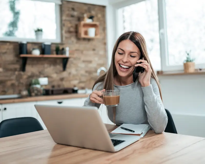 A smiling woman with long brown hair laughs while talking on a mobile phone and holding a glass coffee mug at a wooden kitchen table. A laptop and open notebook with a pen are in front of her, and a rustic brick wall with shelves and plants is visible in the background, evoking the flexibility and ease of working with a virtual assistant from home.