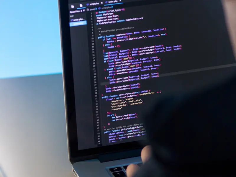 An overhead angle of a laptop screen showing PHP code in a dark IDE, with a developer's hand pointing at the screen. The code includes test functions, class declarations, and parser logic typical of a PHP backend development environment.