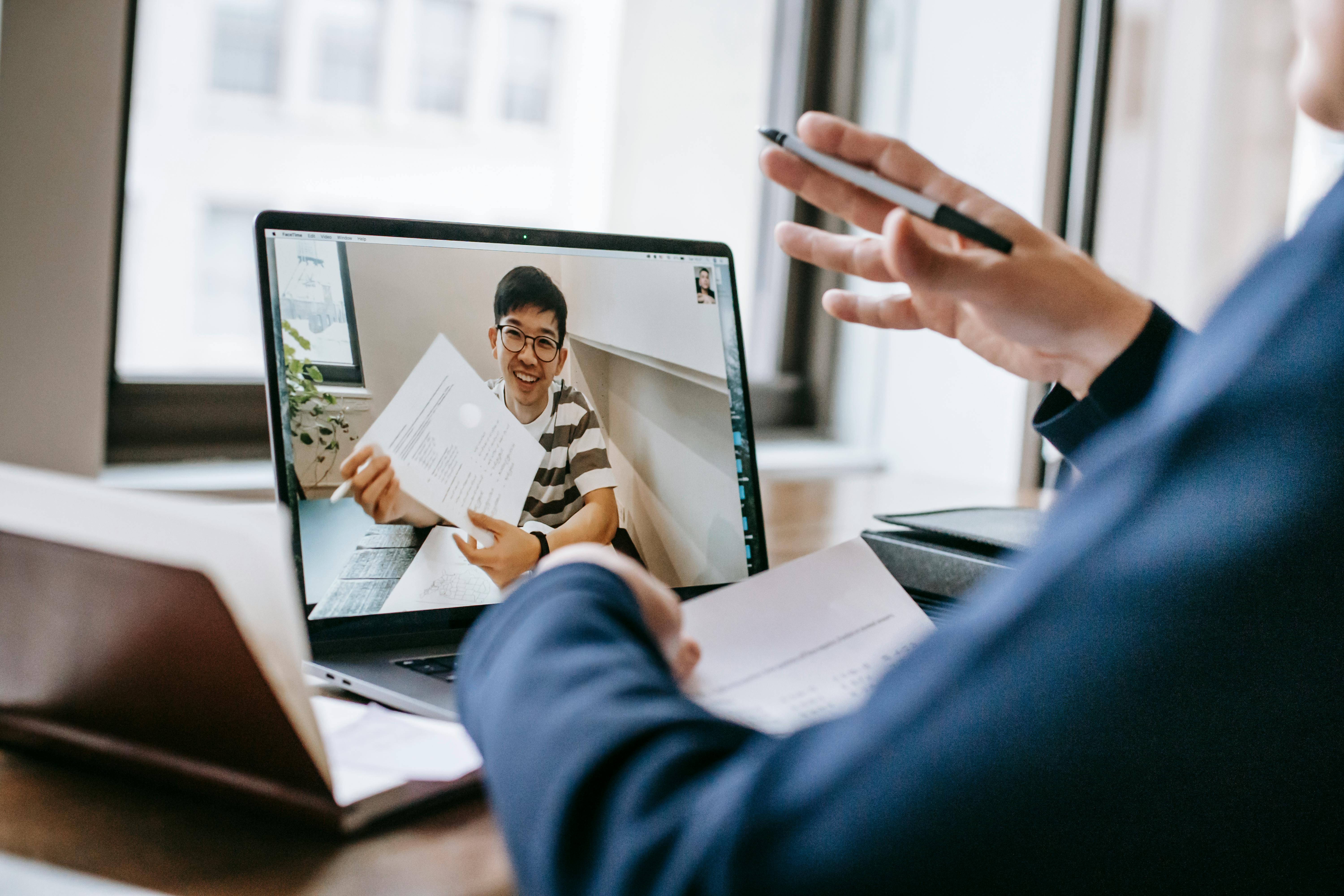 A close-up view from behind a person in a navy suit holding a pen and papers while engaging in a video call. On the laptop screen, a smiling young man in glasses holds up a document, representing a remote business development representative conducting a discovery or proposal meeting with a prospect.