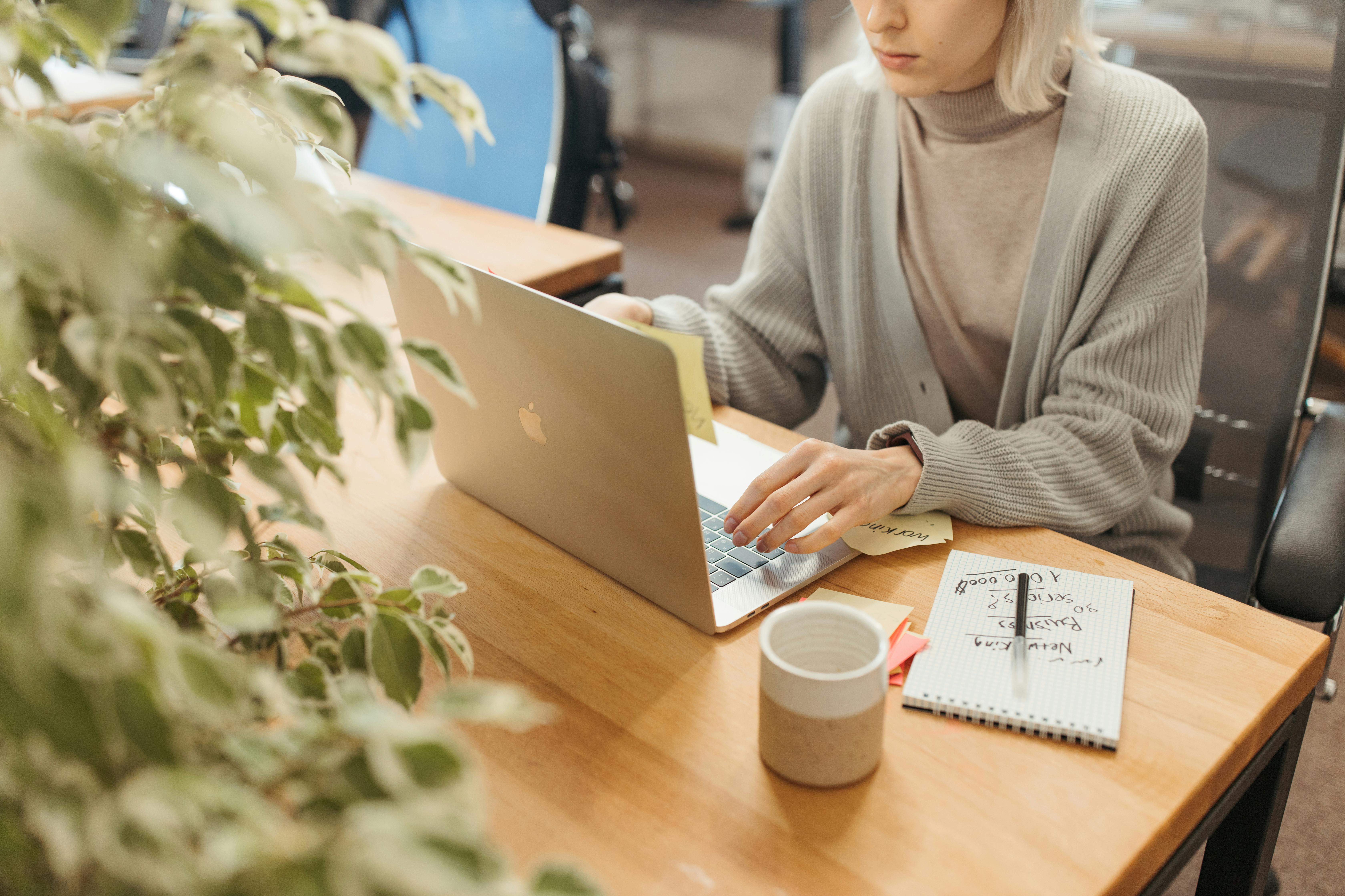 A woman with light hair wearing a grey turtleneck sweater works on a MacBook at a wooden desk in a bright, plant-filled office. A notepad with handwritten notes including words like "networking" and "business" sits beside a mug, depicting a remote business development professional organizing their outreach strategy.