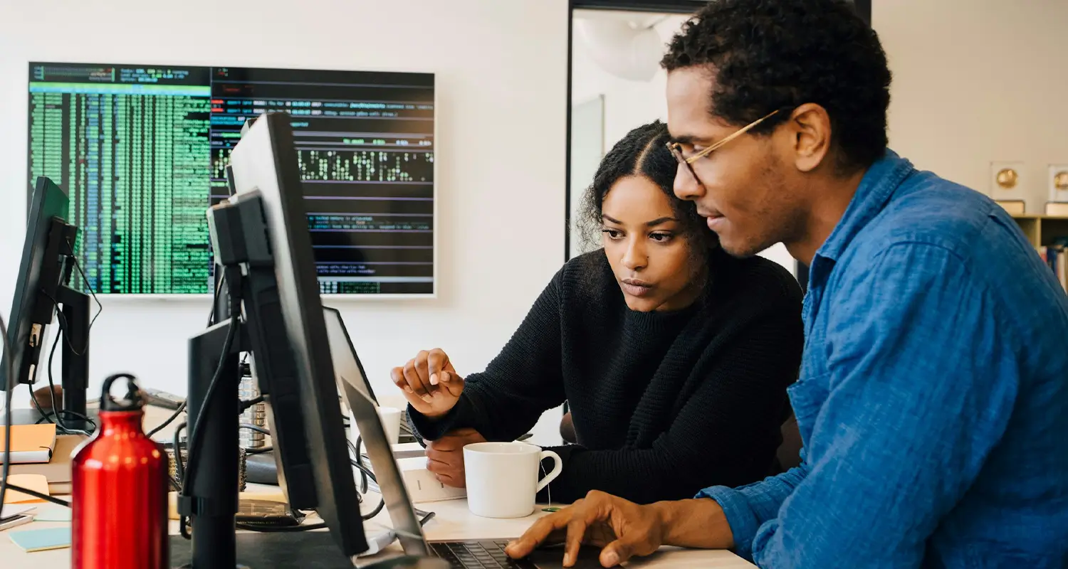 Two data scientists collaborating at a desk with a large monitor showing code, analyzing data together