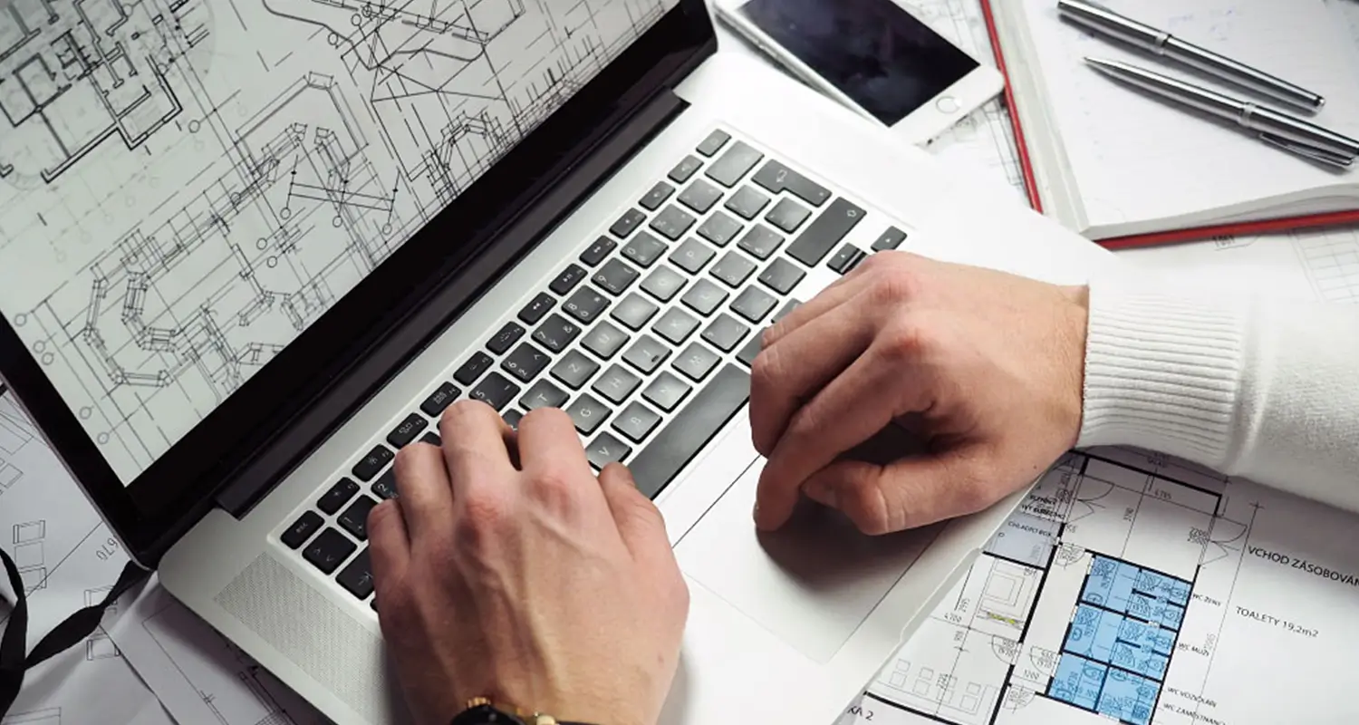 An overhead view of a person's hands typing on a laptop whose screen displays detailed architectural blueprints. A blue-highlighted floor plan, pens, a smartphone, and a notebook are arranged on the white desk surface, depicting a property improvement specialist reviewing technical construction or renovation plans.