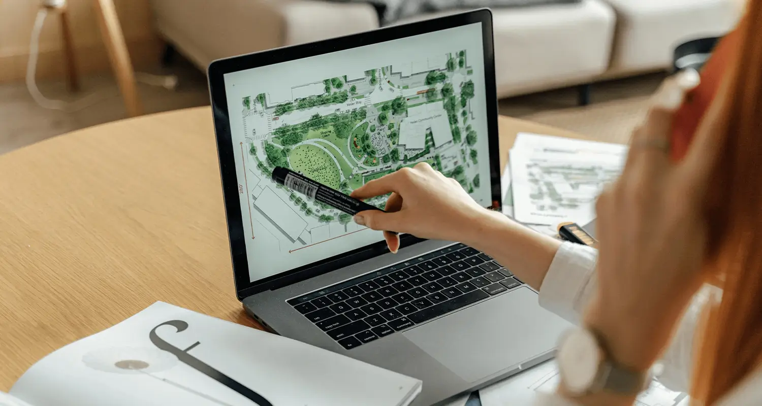A woman with red hair points a marker toward a digital architectural site plan displayed on a laptop screen, showing a bird's-eye layout of a property with green landscaping. Printed documents and plans are spread on the round wooden table beside her, suggesting a property improvement planning session.