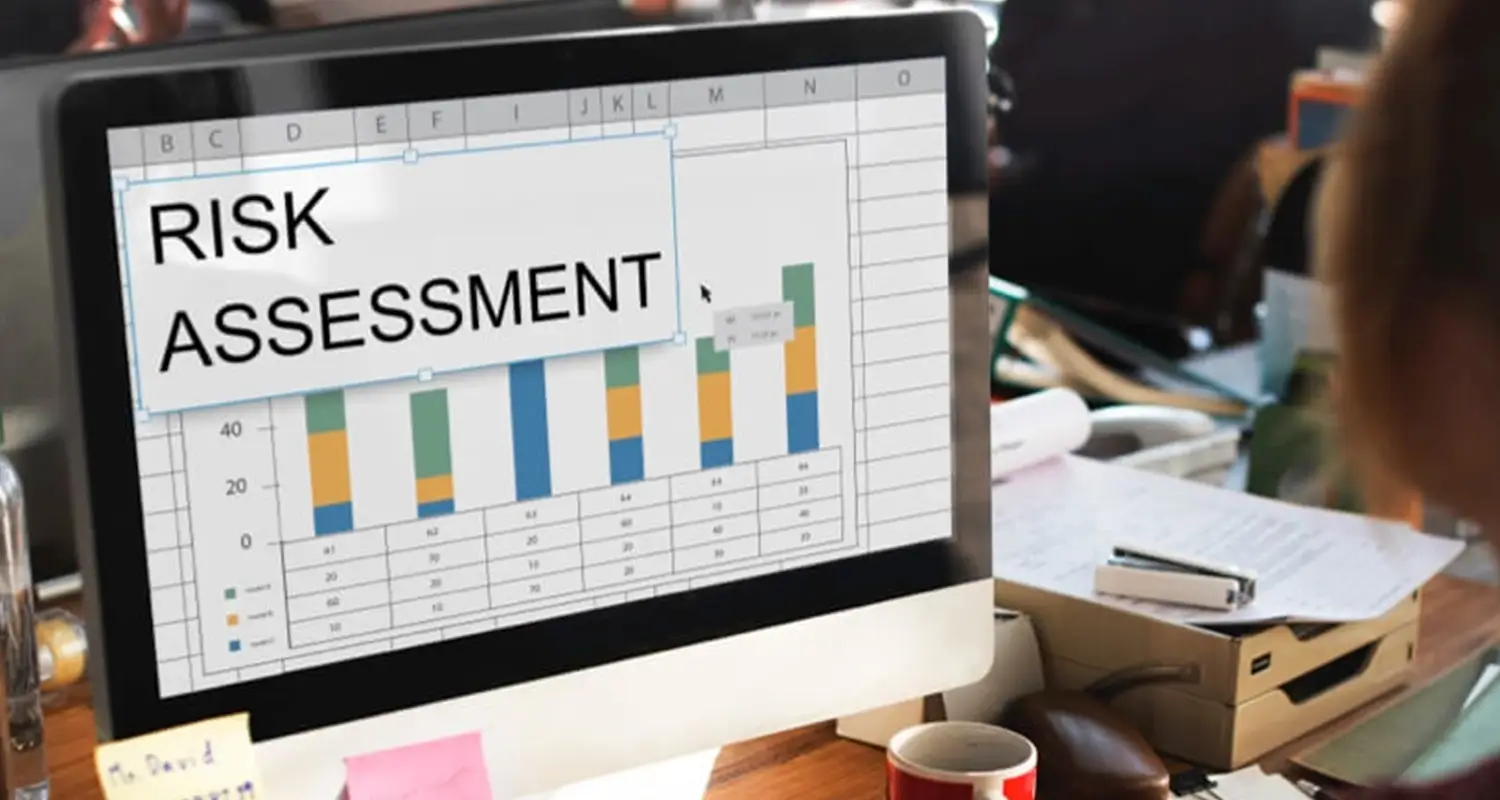 A desktop monitor displays a spreadsheet labeled "Risk Assessment" with a stacked bar chart showing data across multiple columns in blue, green, and orange. A person sits in the background next to stacked paperwork and office materials.