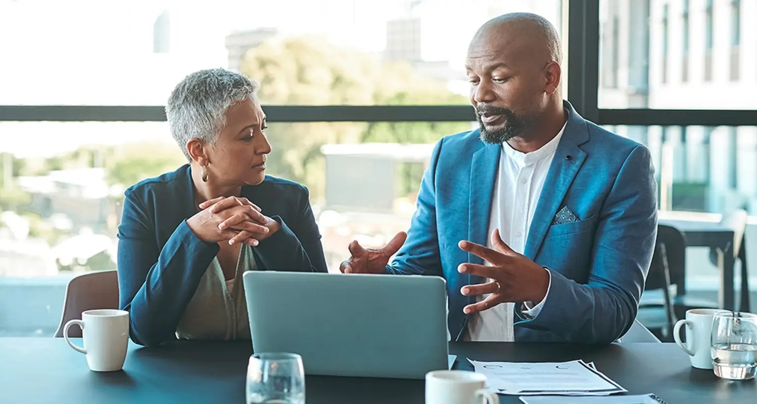 Two business professionals — a woman with short gray hair and a man in a blue blazer — sit at a conference table having a focused discussion in front of an open laptop. Coffee cups and documents are visible on the table, with a city view through floor-to-ceiling windows in the background.