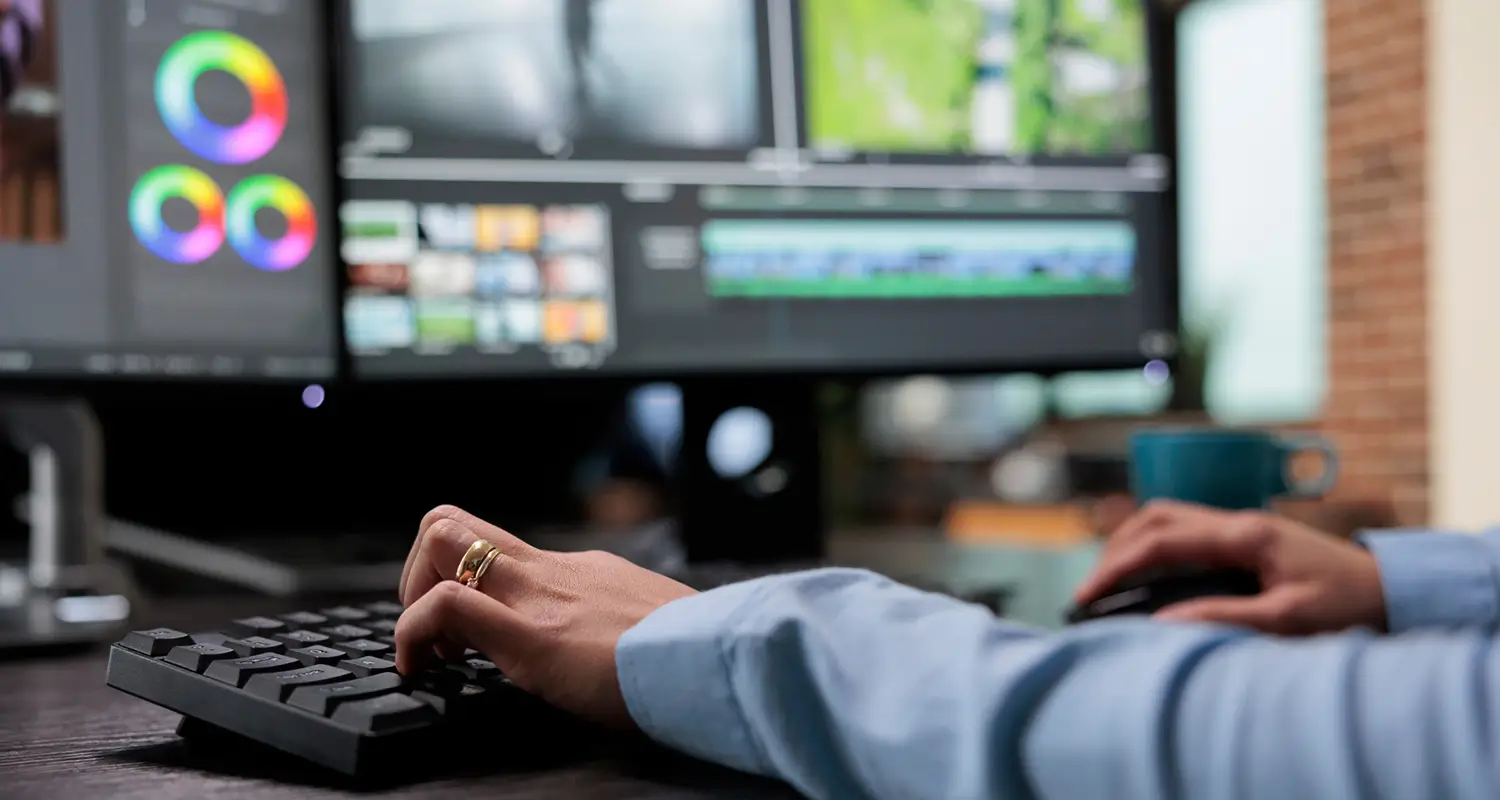 Close-up of a video editor's hands typing on a black keyboard, with dual monitors in the background showing a video editing timeline with color grading wheels and multi-track footage clips in a professional editing suite.