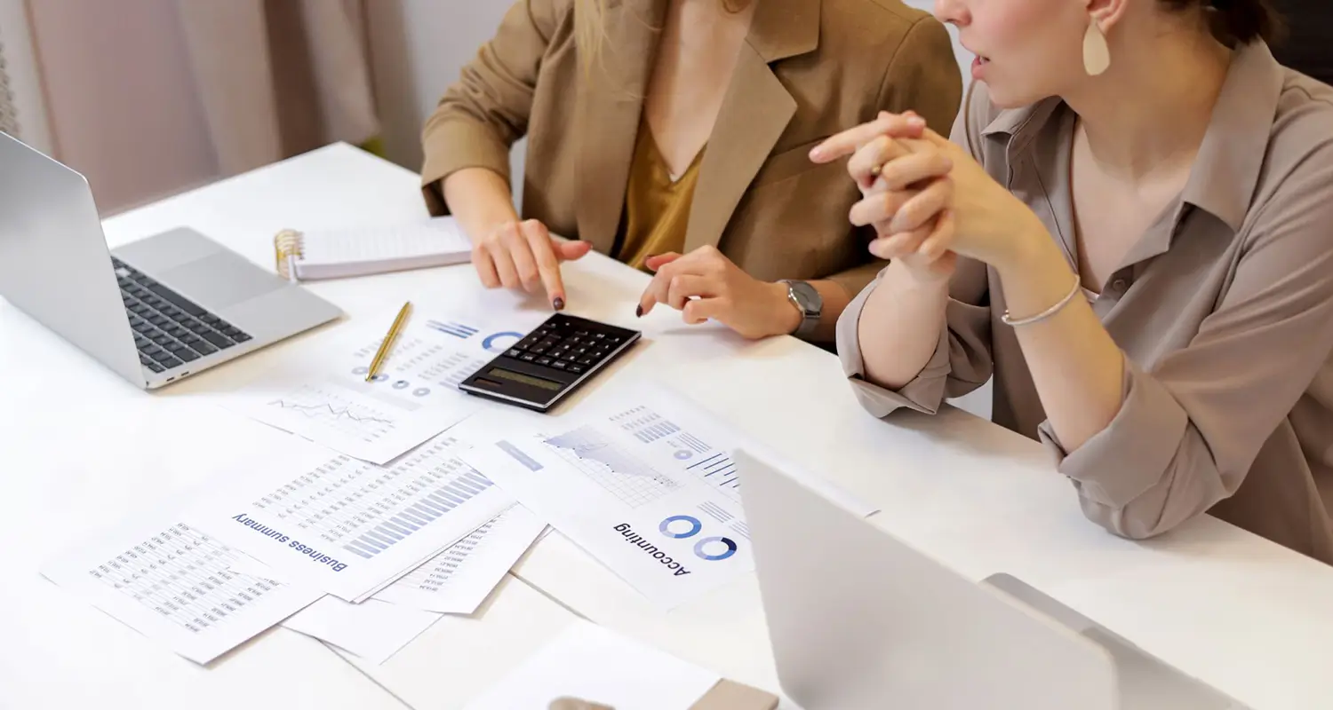Two businesswomen reviewing accounting and business summary documents spread on a desk, with a calculator and laptop, discussing financial data