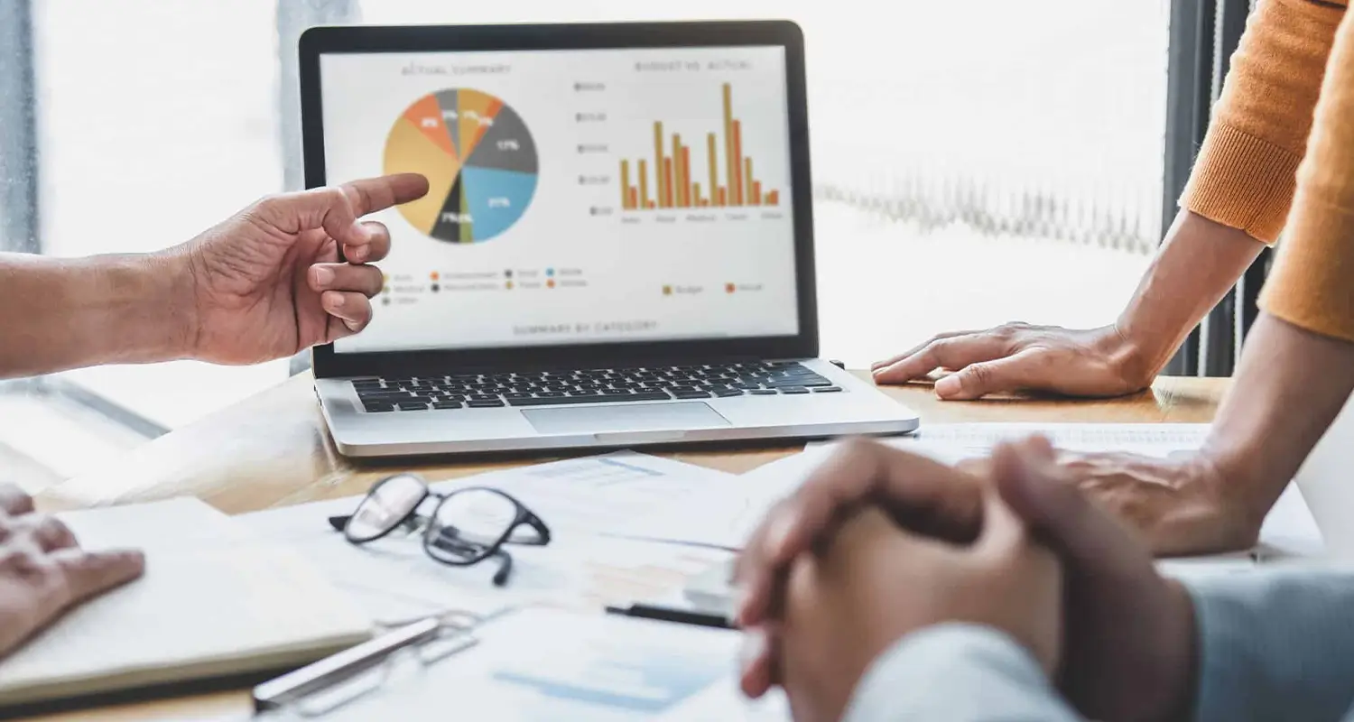 Two people sit across from each other at a meeting table as a third person points at a laptop screen showing a pie chart and bar graph financial dashboard. Printed reports and glasses are spread on the table, representing a workforce planning session where US business leaders analyze data to inform global hiring decisions.