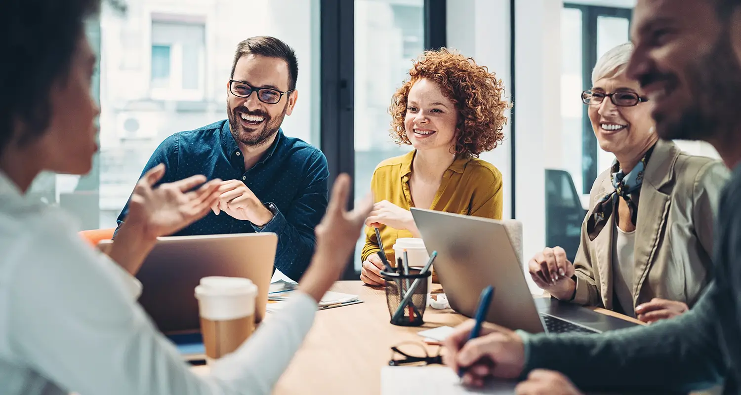 A diverse group of five business professionals sit around a conference table laughing and engaged in a lively discussion, with laptops, tablets, coffee cups, and notebooks in front of them. The energetic atmosphere reflects the collaborative workforce planning conversations that drive strategic global hiring decisions at US growth-stage companies.
