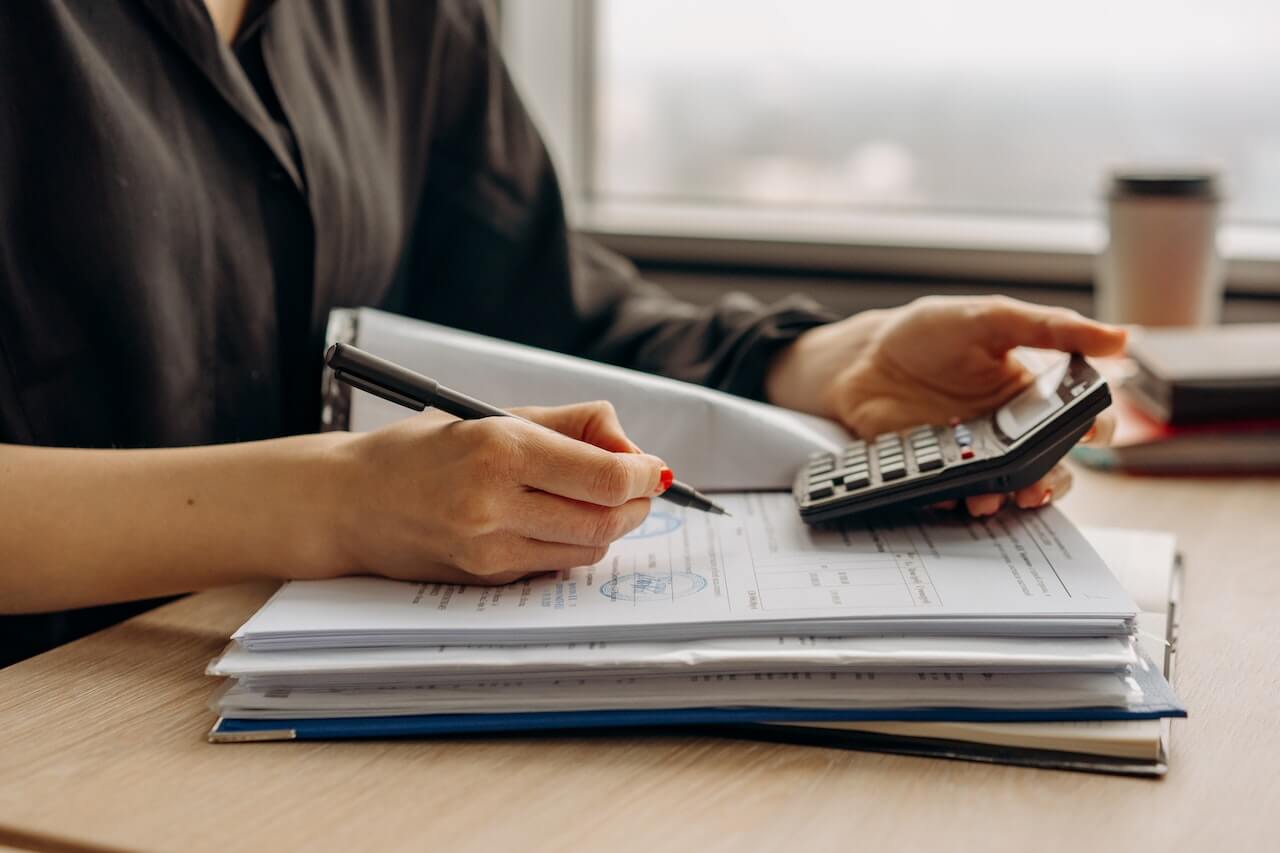 Person writing on financial documents while using a calculator, with stacked file folders on a desk