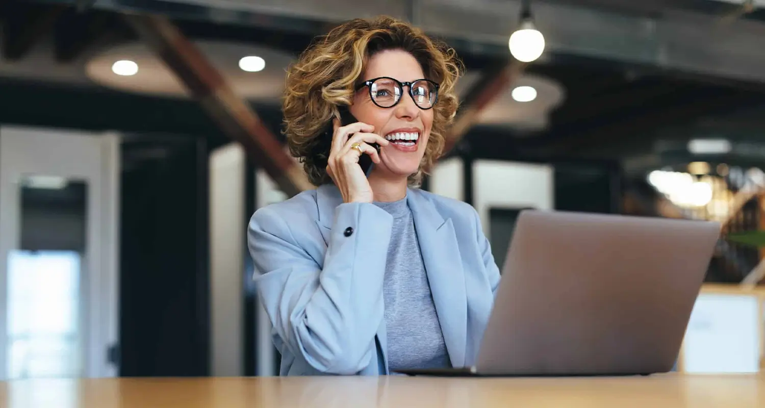A confident woman with short curly hair, glasses, and a light blue blazer laughs while talking on her mobile phone, with a laptop open in front of her in a modern open office. She represents an energetic offshore sales representative naturally building rapport with clients and closing deals remotely.