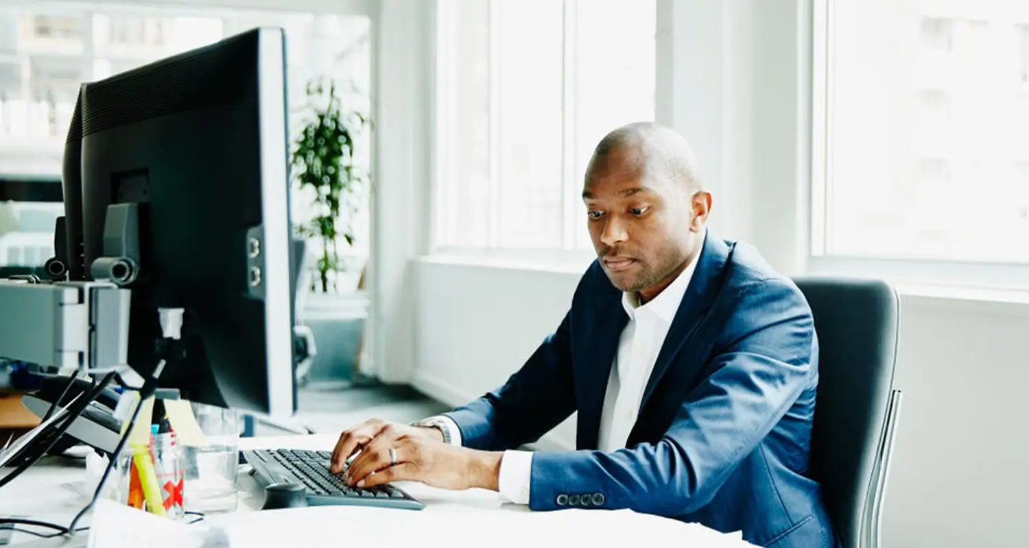 A Black man in a blue suit jacket sits upright at a bright office desk, typing on a keyboard in front of a large monitor. His focused, professional demeanor represents a high-performing offshore sales representative who brings discipline and drive to a remote sales role, delivering results regardless of geographic location.