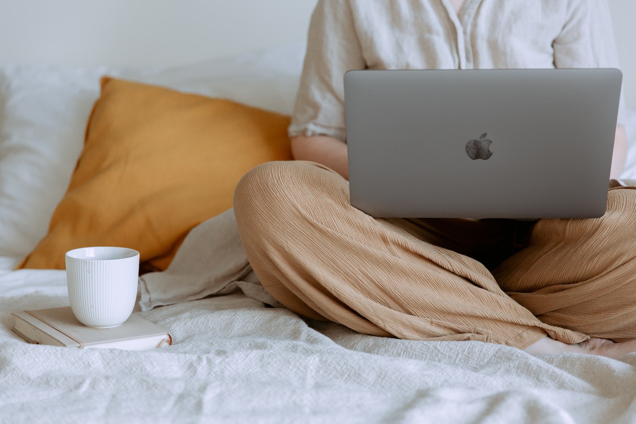 A person's lower body shown seated cross-legged on a white bed with a gray Apple MacBook laptop resting on their lap, next to an orange throw pillow and a white coffee mug on a notebook. The image depicts a remote worker or executive assistant working from a comfortable home setup.