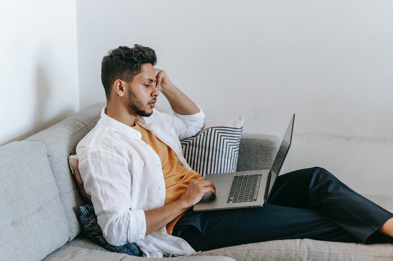 A stressed-looking man sits on a gray couch, head in his hand, working on a laptop in a modern apartment. The image represents an overwhelmed executive who could benefit from delegating calendar management to a virtual assistant.