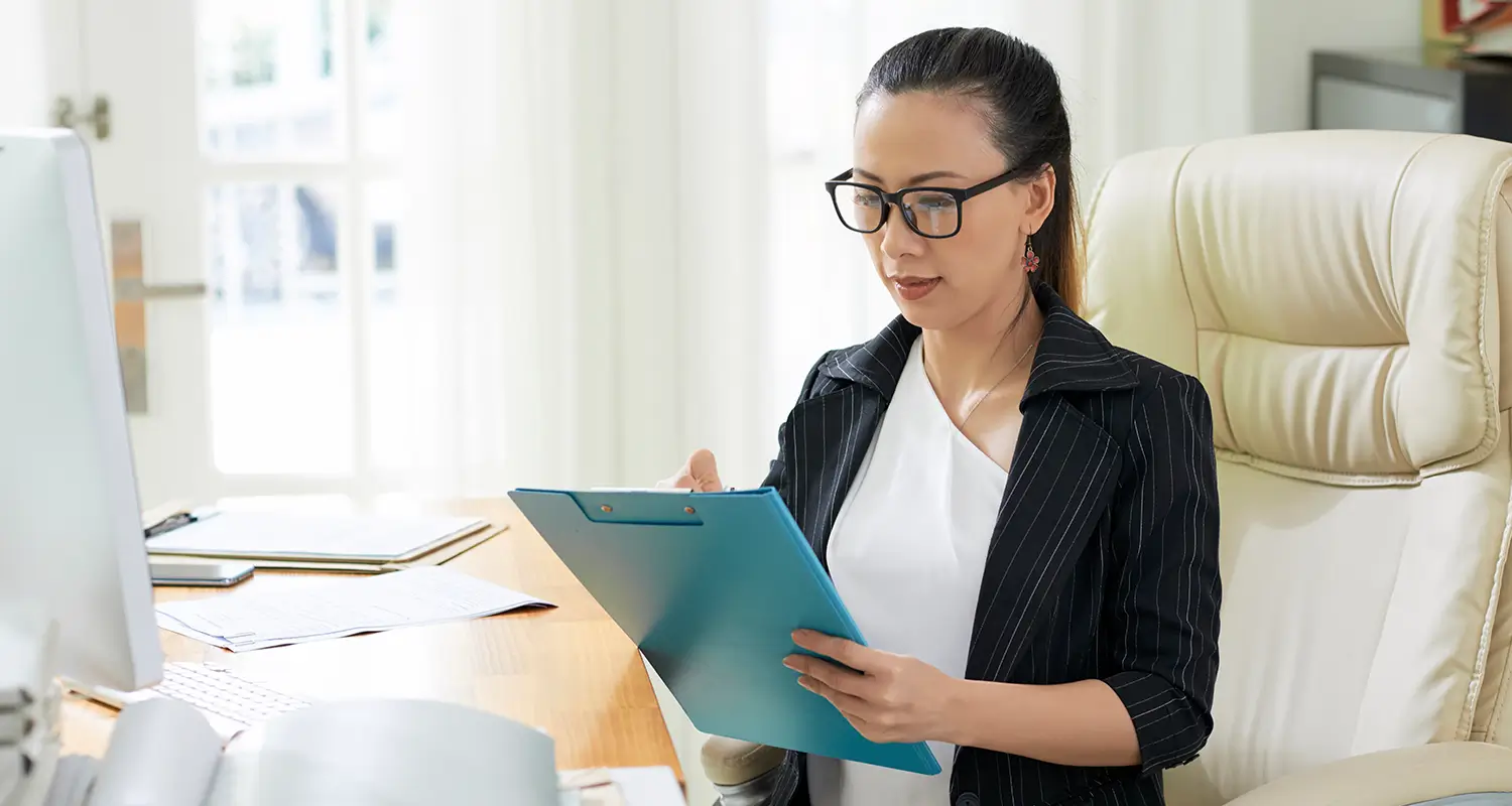 Female procurement manager in glasses reviewing a clipboard document at her office desk
