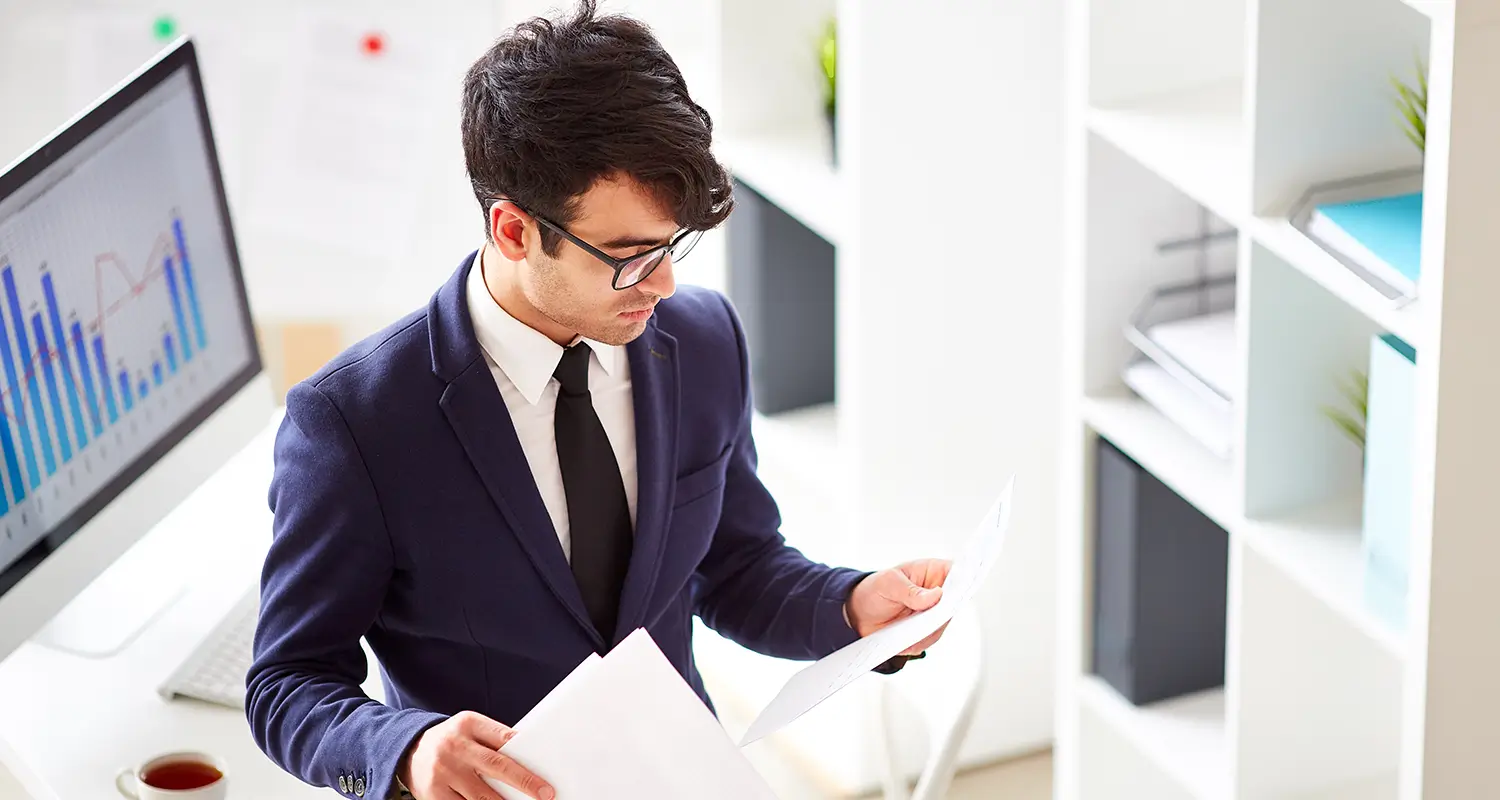 Procurement specialist in a suit reviewing documents with a financial chart on screen behind him