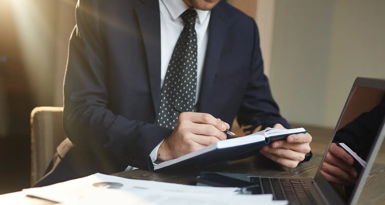 Attorney in a suit writing notes in a notebook with documents and a laptop on the desk