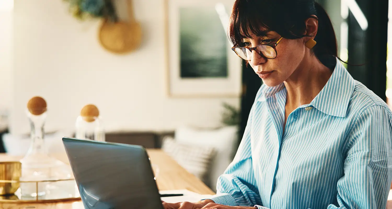 Woman in glasses and a blue striped shirt focused on typing on a laptop at home